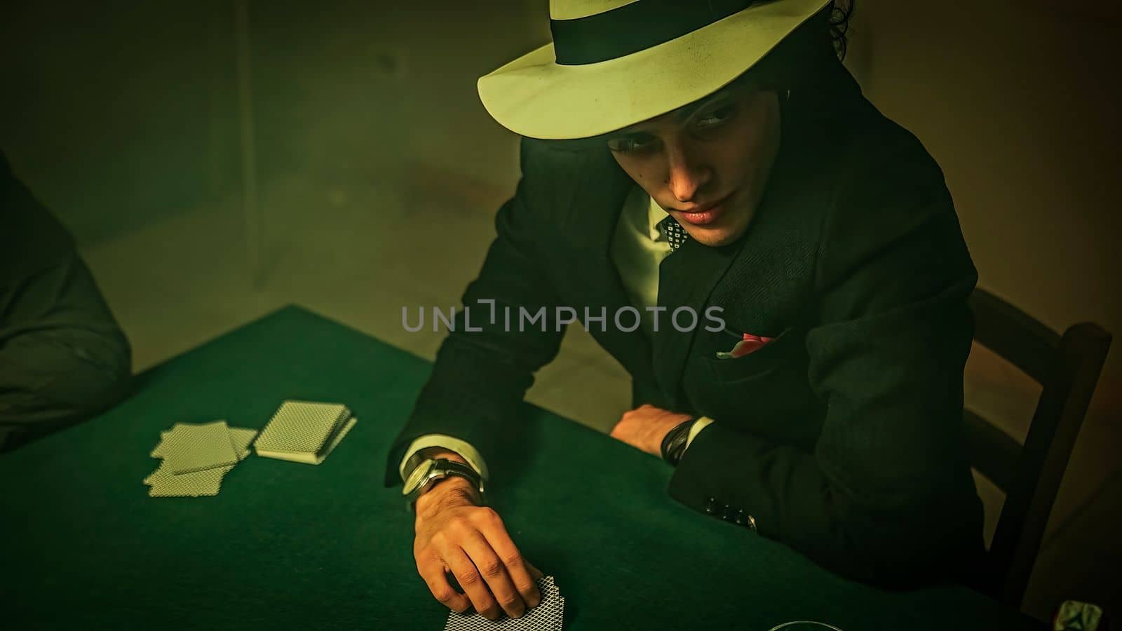 A group of friends are gathered around a green, textured poker table, where a young man dressed in a suit is actively engaged in a game of poker. There is a drink glass visible on the table.