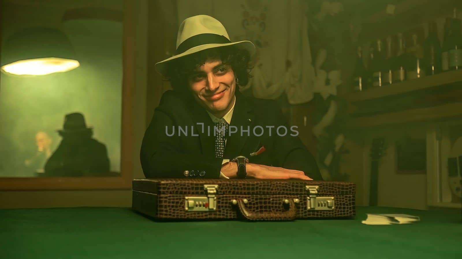 A man sitting at a table in a dimly lit room filled with smoke. He accepts a briefcase with a smile on his face. The scene suggests a negotiation or possibly a shady deal.