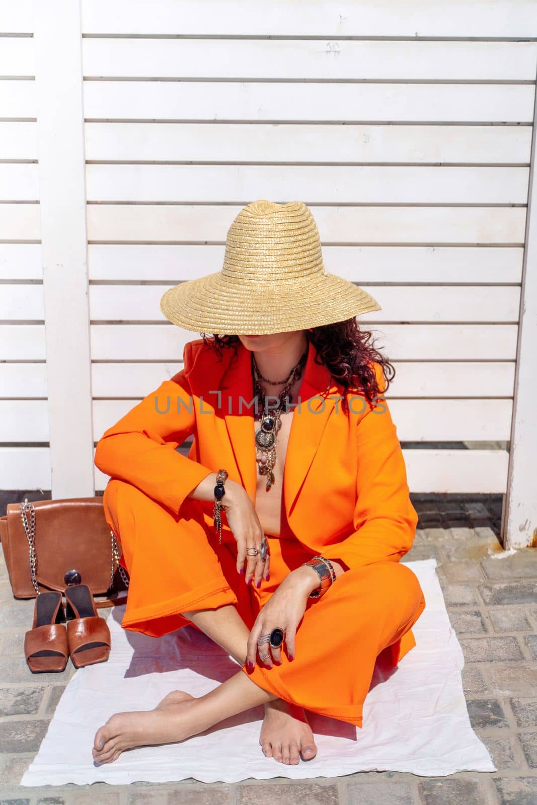 Stylish woman in an orange suit with a hat sits on a rug on a white striped background. On the hands are jewelry rings and bracelets, sandals and a bag stand side by side