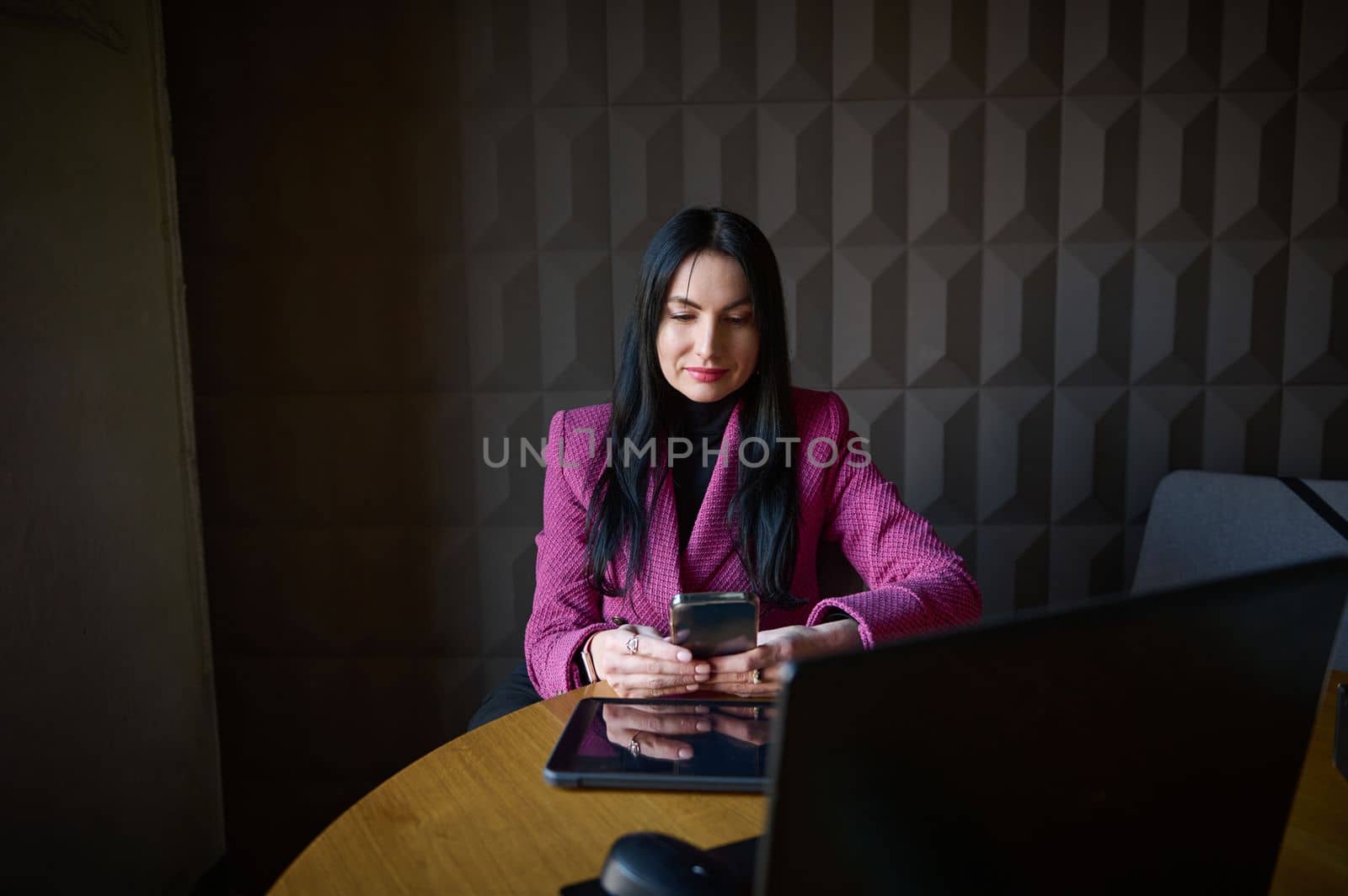 Elegant stylish middle-aged Caucasian woman, successful business lady typing text on her smartphone while telecommuting on laptop from a coworking. Online business. Remote work. People concept