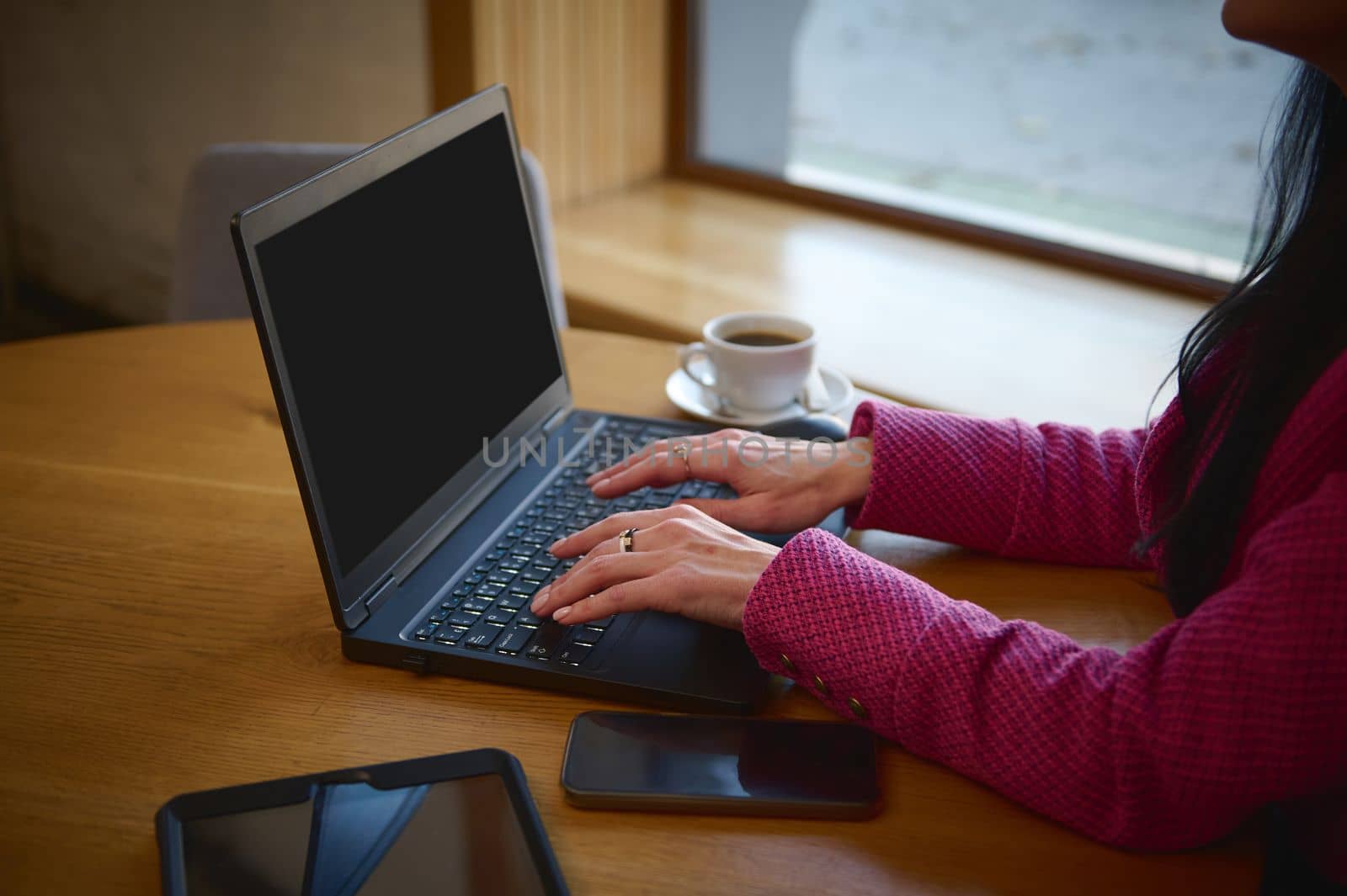 Selective focus on hands of a businesswoman, dressed in bright magenta jacket, on laptop keyboard while telecommuting, sitting at table with a cup of coffee in a cozy cafeteria. Remote work. Business
