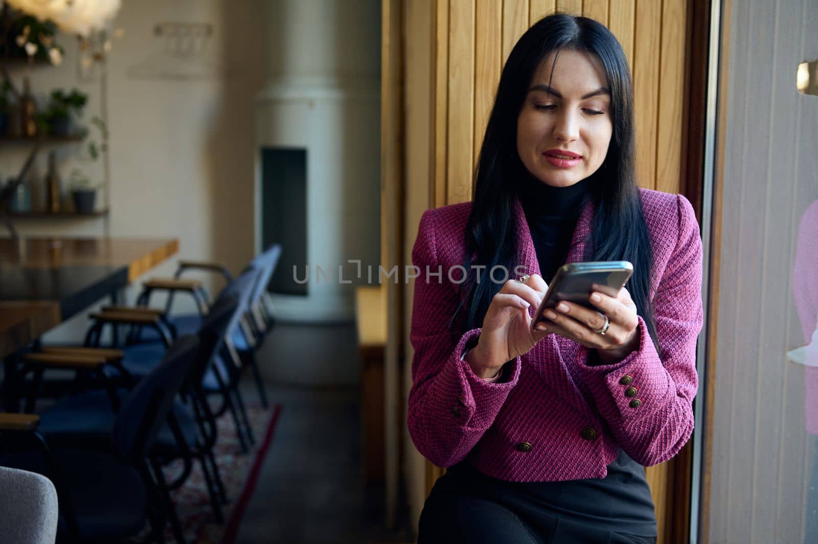 Confident elegant dark-haired Caucasian pretty woman 40s, sitting on the window sill in a meeting room, chatting on her modern smartphone, planning meeting with business partners, investors, employees