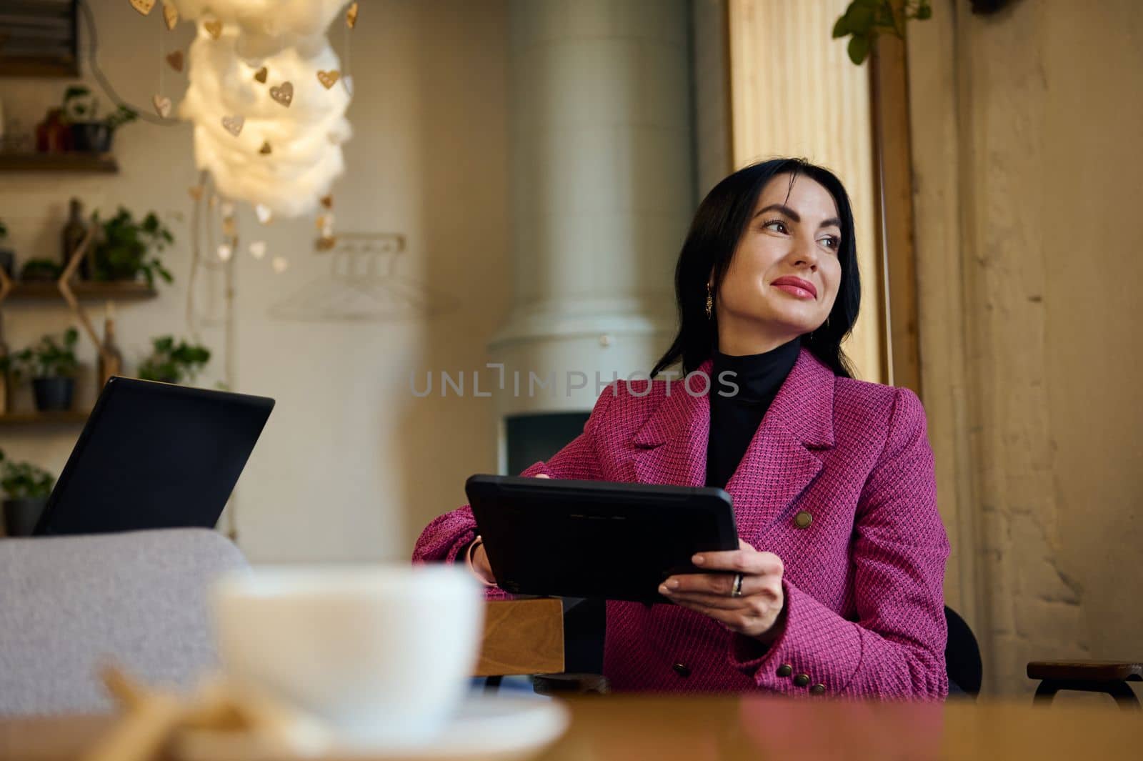 Middle-aged Caucasian confident, successful gorgeous woman, wearing stylish purple casual jacket, working on a digital tablet, waiting for business partners while a coffee break in a cozy restaurant