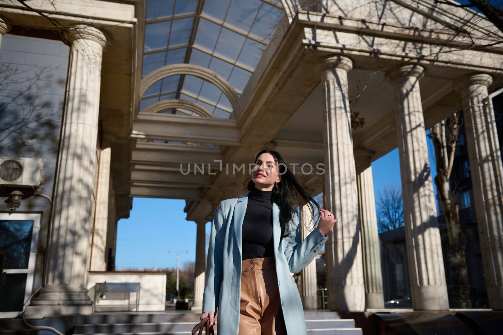 Beautiful dark-haired Caucasian middle-aged woman in casual wear, cheerfully smiling at camera while walking down the urban street on a beautiful sunny early spring day. People. Urban. life Lifestyle