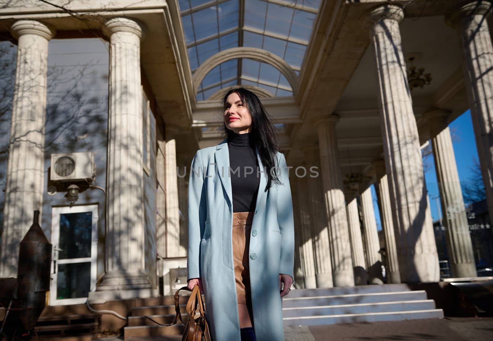 Caucasian elegant stylish dark-haired middle aged pretty woman in stylish blue coat, holding handbag, enjoying morning walk on the city street on a sunny early spring day. People and lifestyle concept