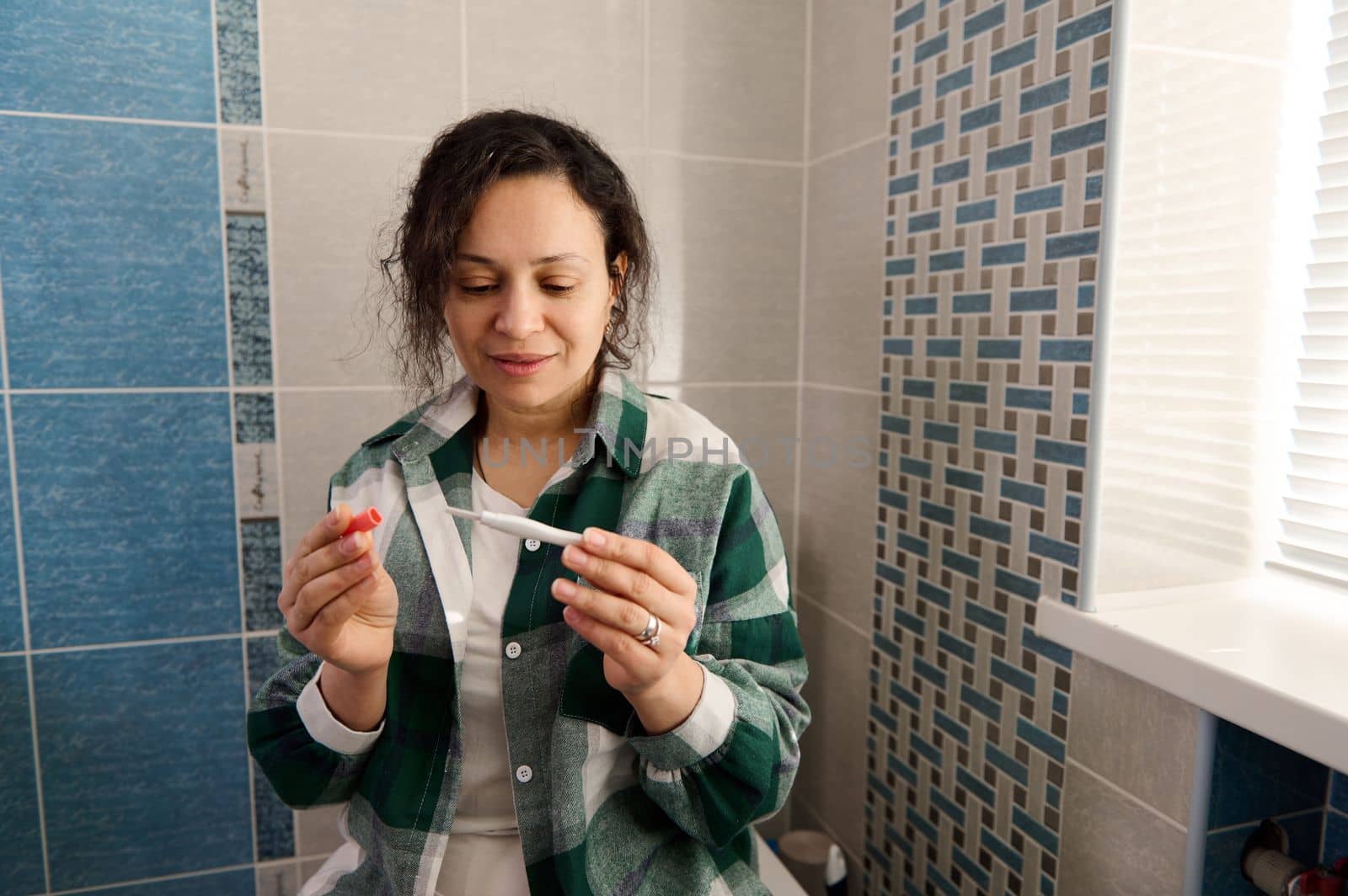 Multi-ethnic middle-aged woman in green checkered shirt, sitting on the toilet bowl in the home bathroom, taking pregnancy test, awaiting the result. People, lifestyle, maternity and pregnancy concept