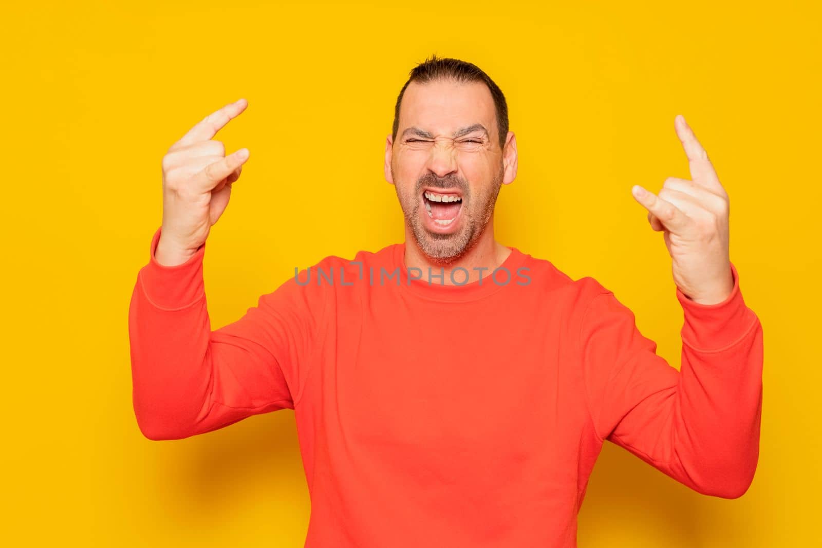 Rock fan gestures. Caucasian bearded man in red jumper doing rock symbol with hands up while standing against orange background. body language concept