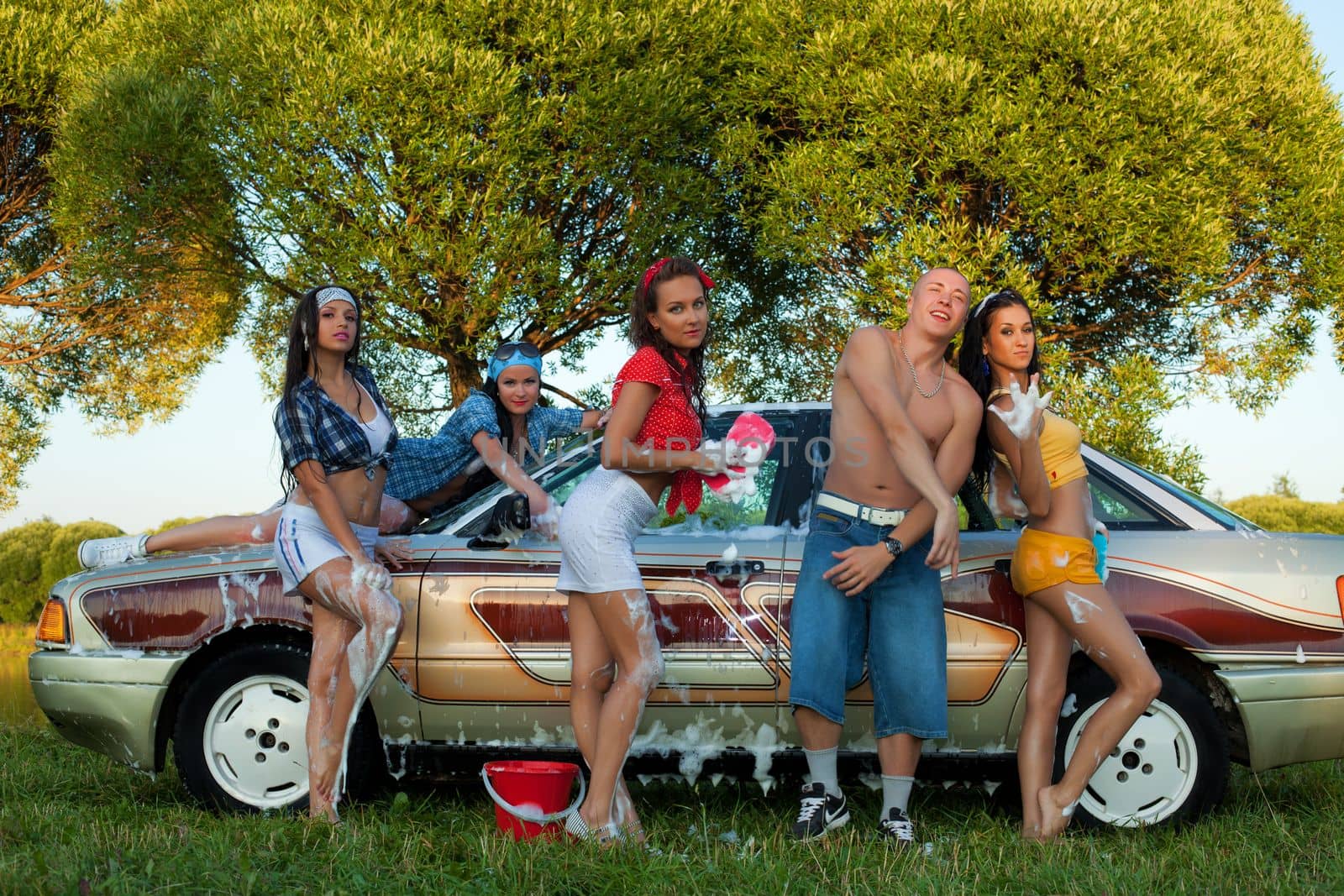 Cheerful female friends sitting on trunk of vintage car and looking at camera