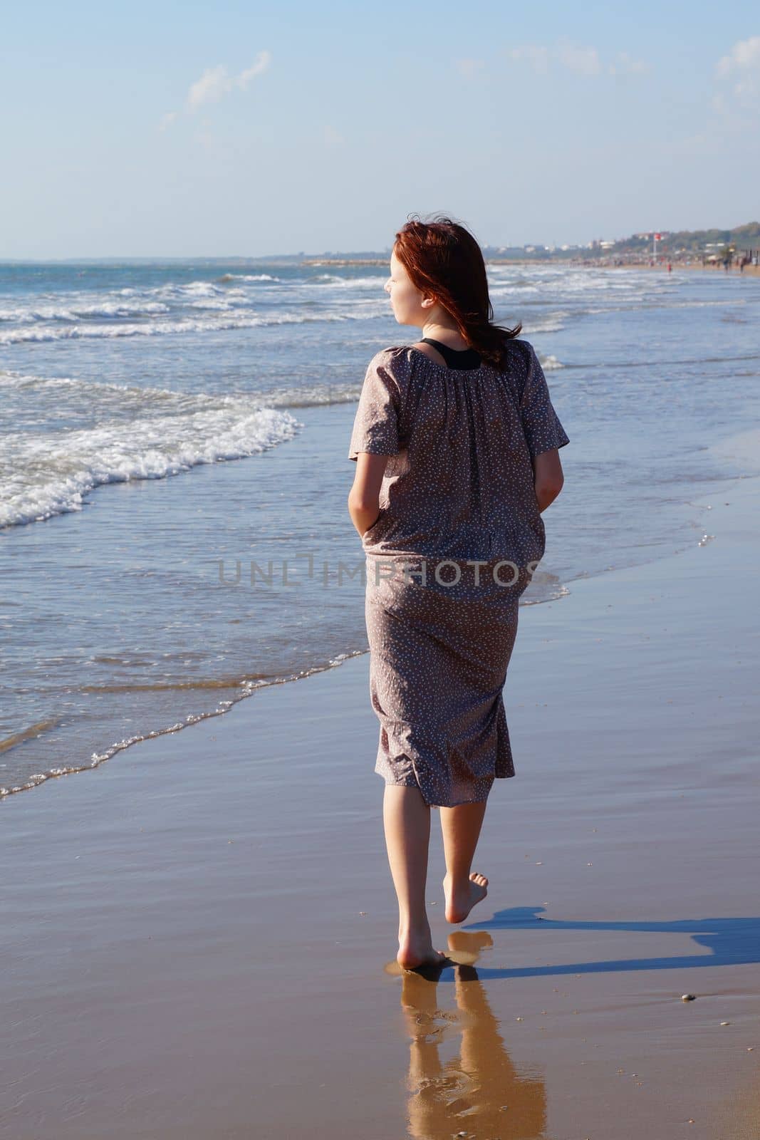 a red-haired barefoot teenage girl in a dress walks along the seashore along the water's edge, back view