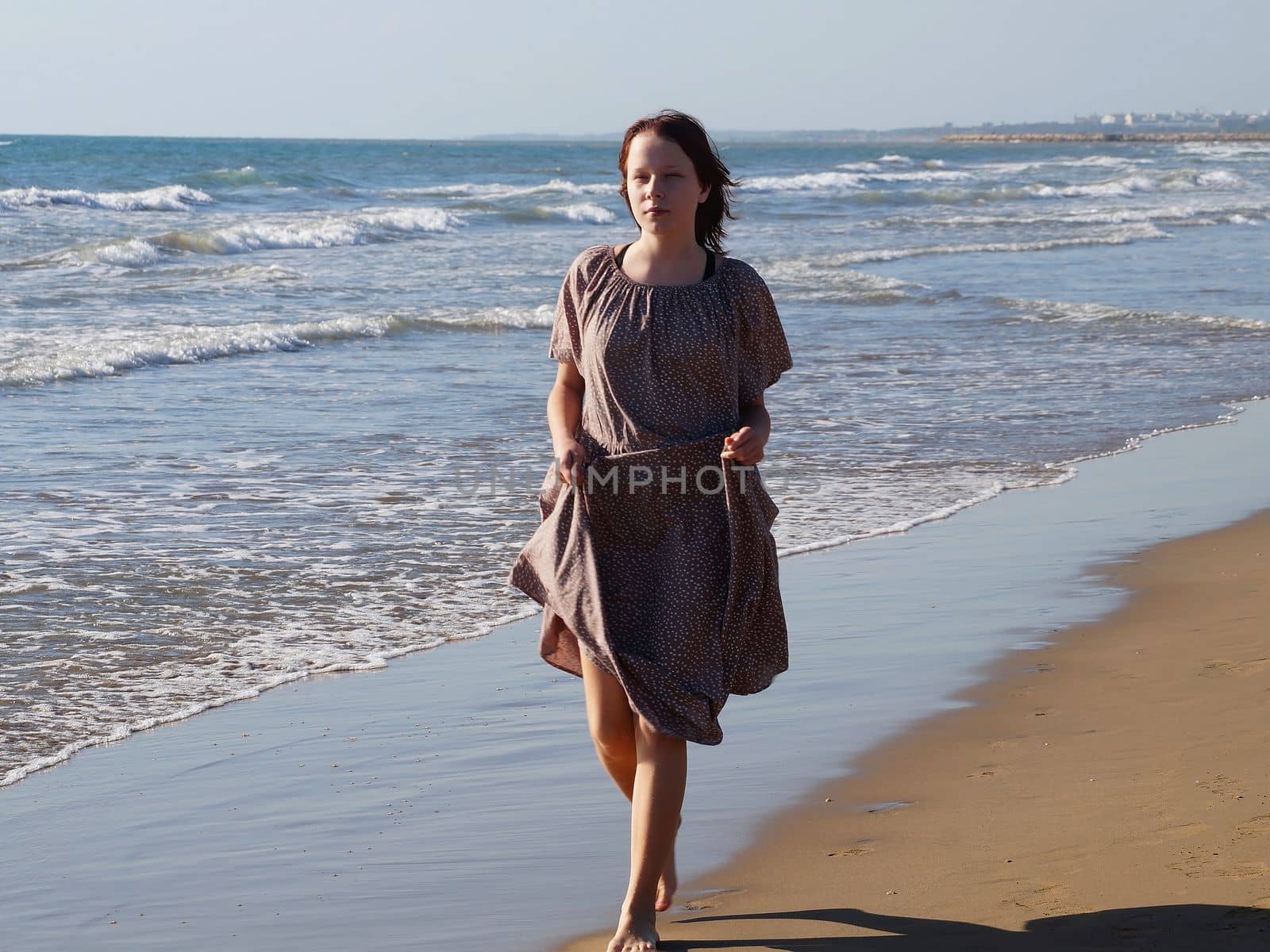 a red-haired barefoot teenage girl in a dress walks along the seashore along the water's edge.