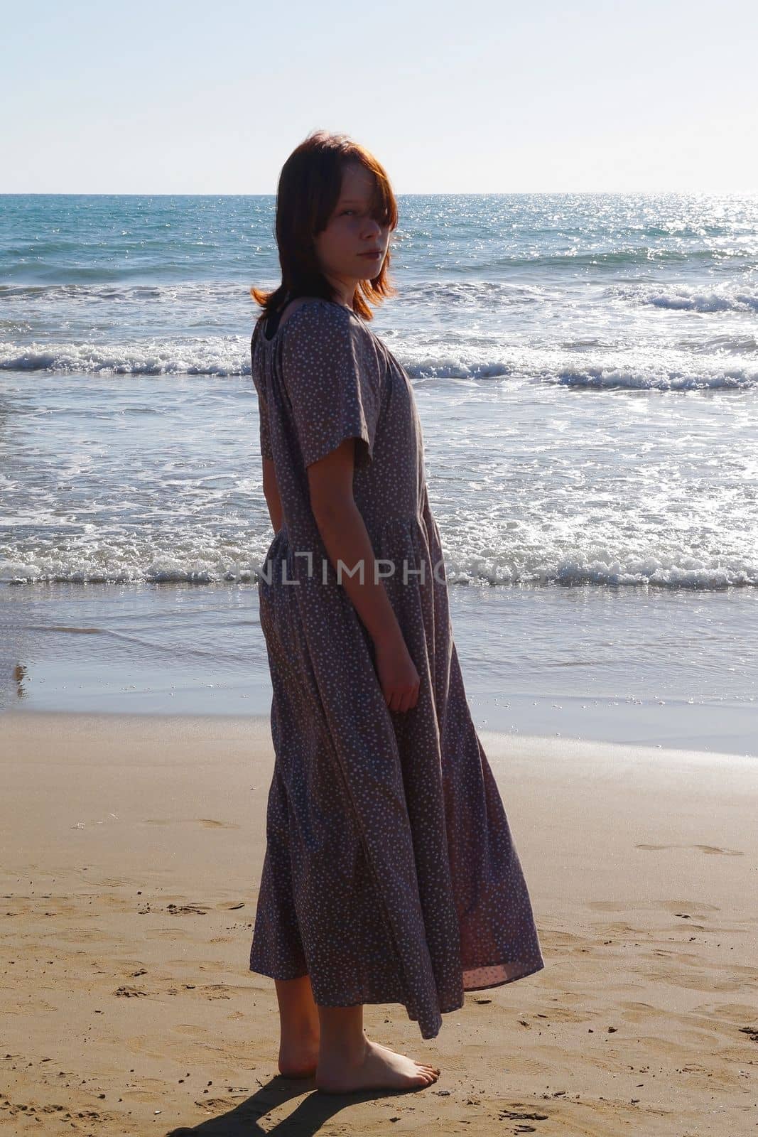 barefoot red-haired teenage girl in a long dress on the sand at the water's edge.