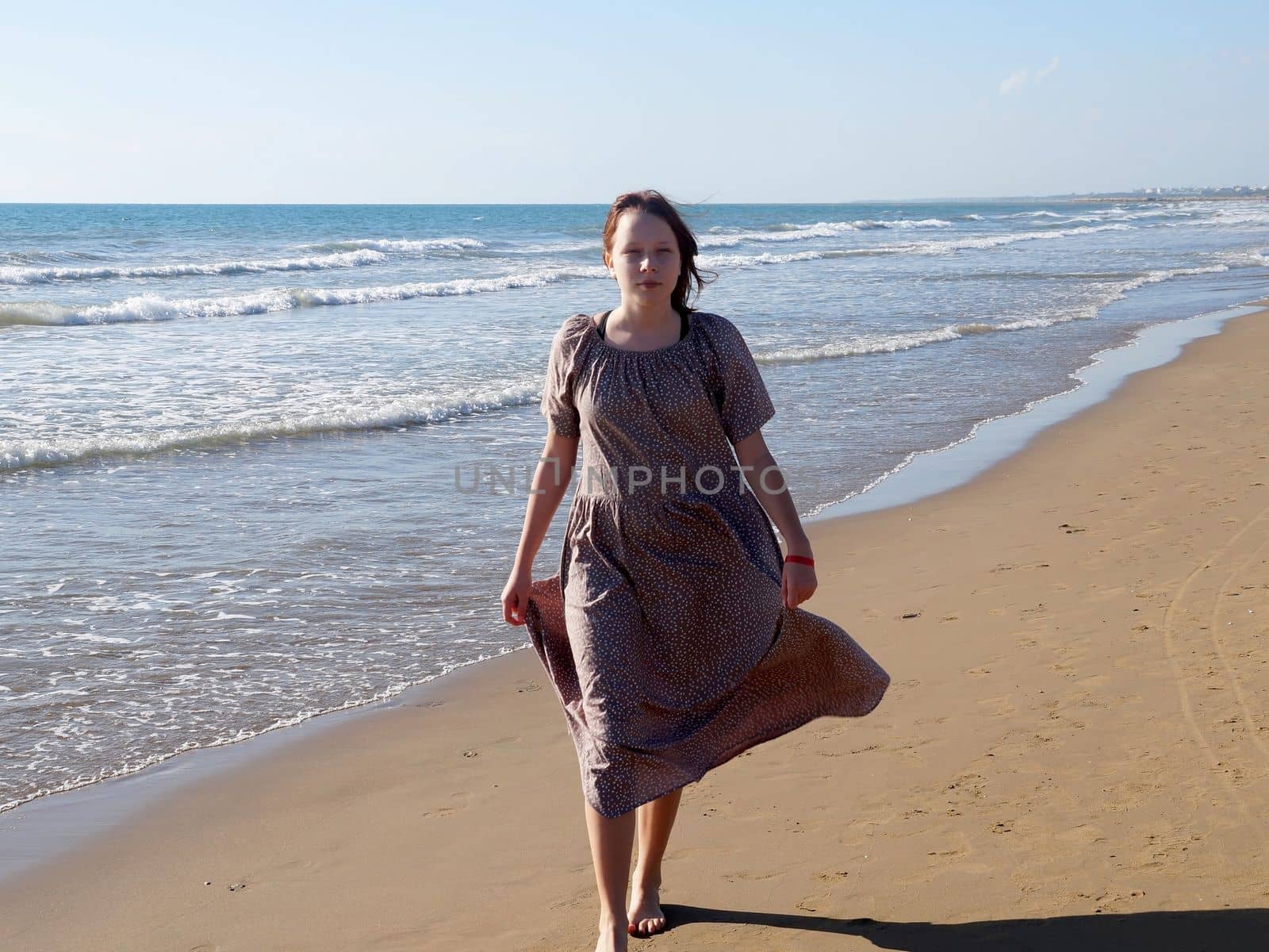 a red-haired barefoot teenage girl in a flowing dress walks along the seashore along the water's edge