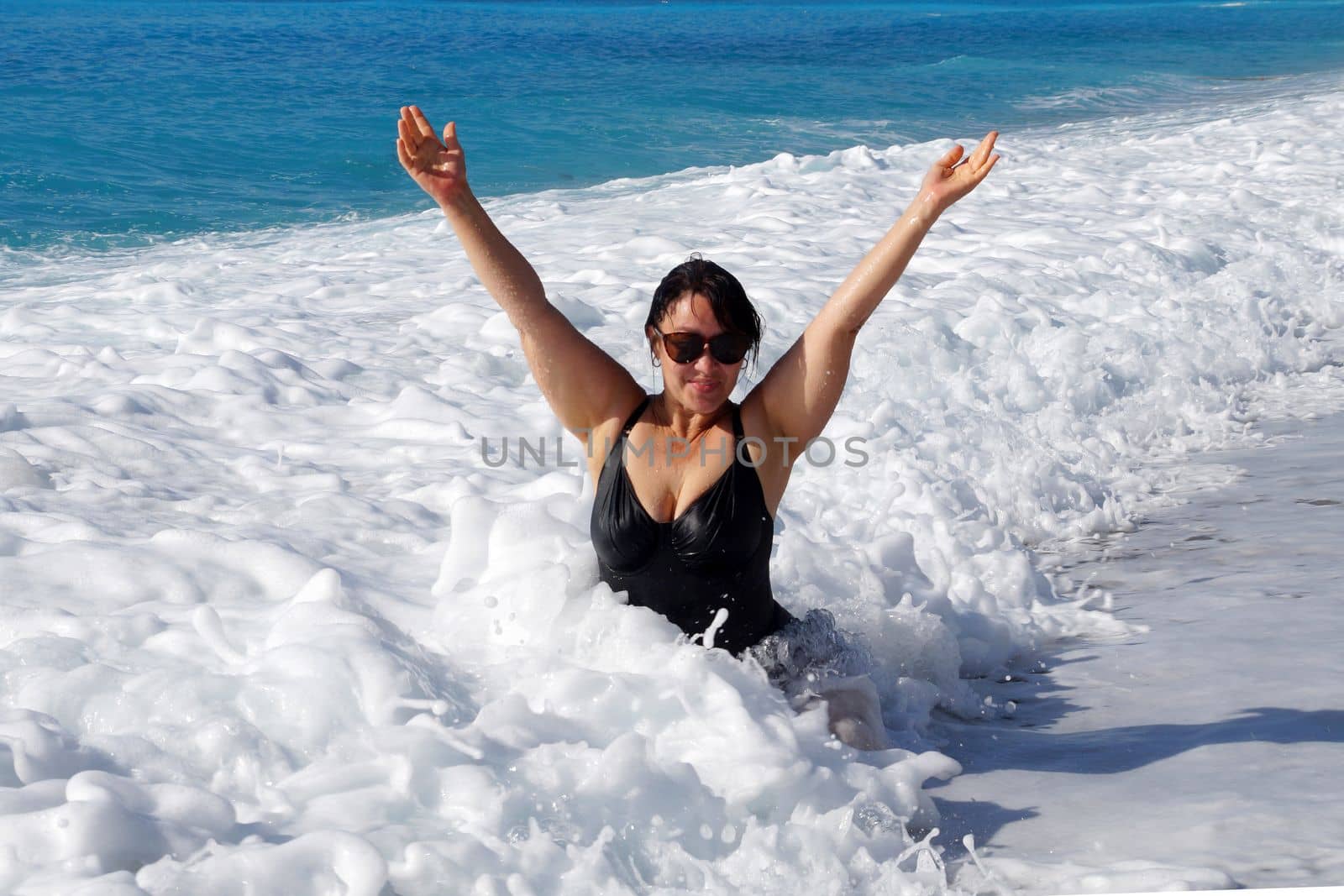 smiling curvy brunette in a black swimsuit sits in the sea with her hands up by Annado