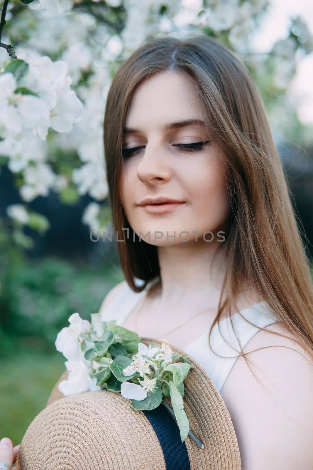 Beautiful young girl in white dress and hat in blooming Apple orchard. Blooming Apple trees with white flowers