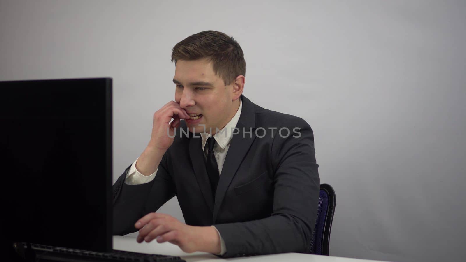 A young businessman at the workplace bites his nails from nerves. IT specialist in a suit is nervous while sitting in the office and looking at the PC monitor. 4k