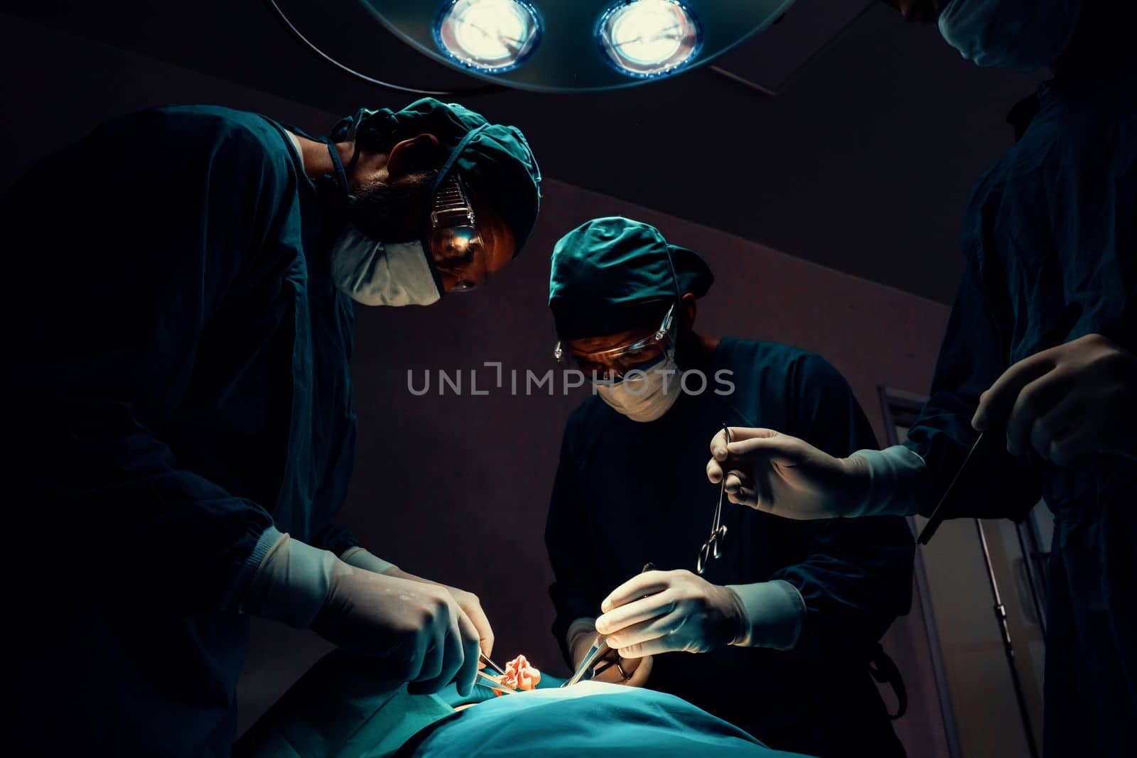 Surgical team performing surgery to patient in sterile operating room. In a surgery room lit by a lamp, a professional and confident surgical team provides medical care to an unconscious patient.