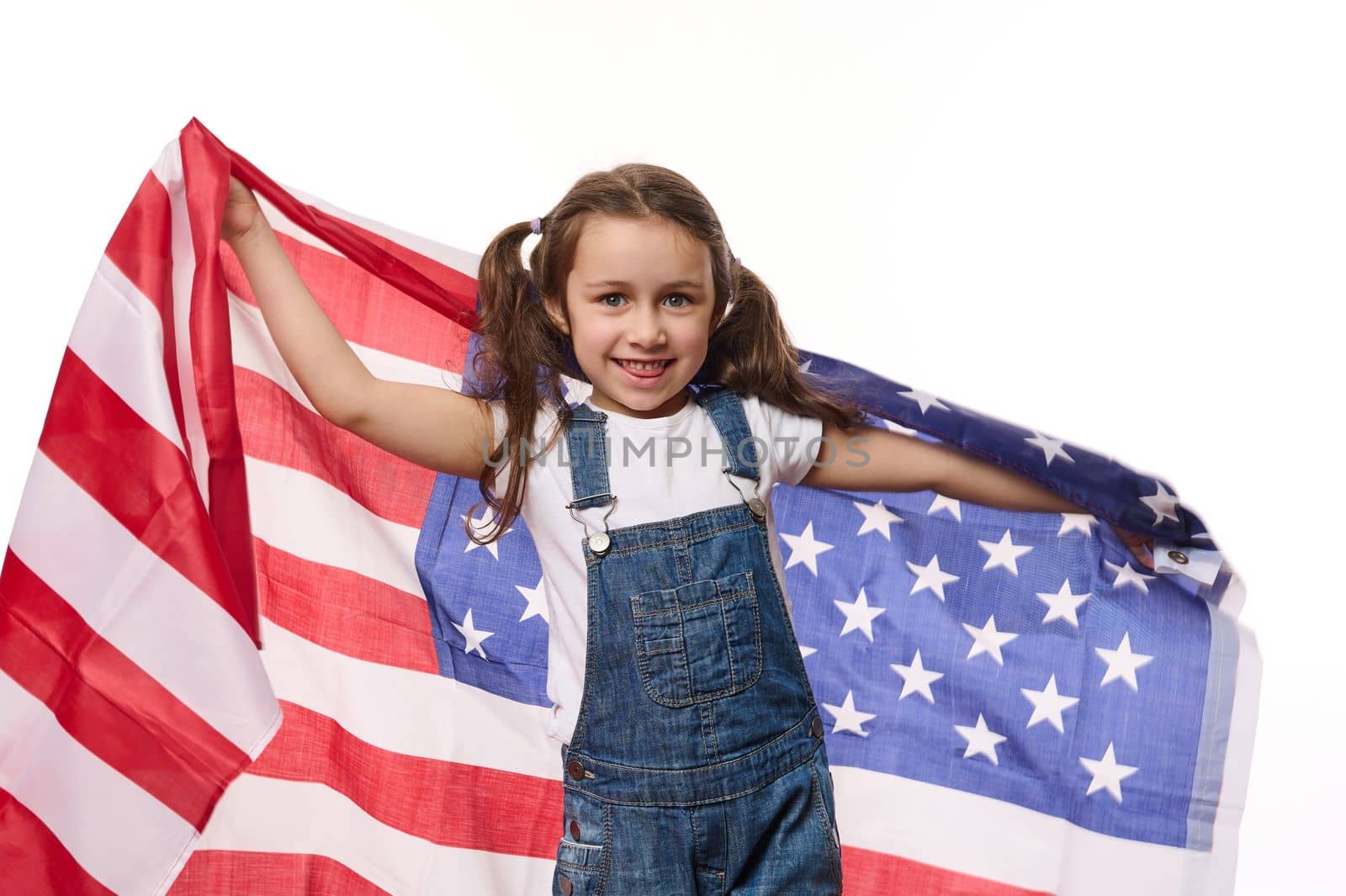 Adorable American child girl in blue denim overalls, carries USA flag, celebrates Independence Day on July 4th. Citizenship, immigration, emigration, winning the green card lottery and freedom concept