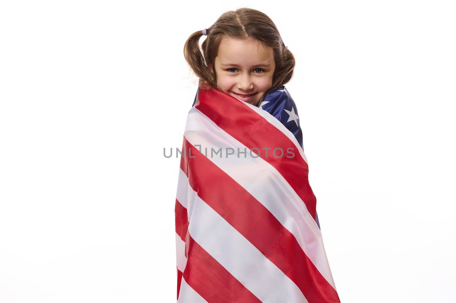 Beautiful patriotic American citizen, a lovely little child girl with two ponytails, covered in USA flag on Independance Day - 4th July, cutely smiles looking at camera, isolated over white background