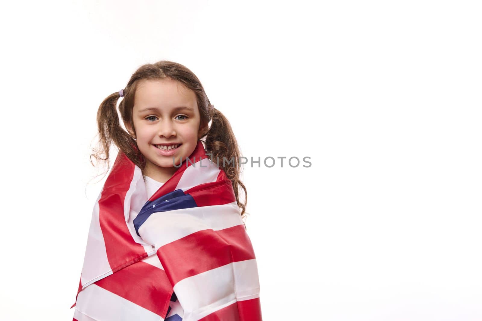 Lovely preschooler girl with two cute ponytails, wrapping in USA flag, celebrating the independence Day of United States of America on July 4 th. Concept of human rights and freedoms. White background