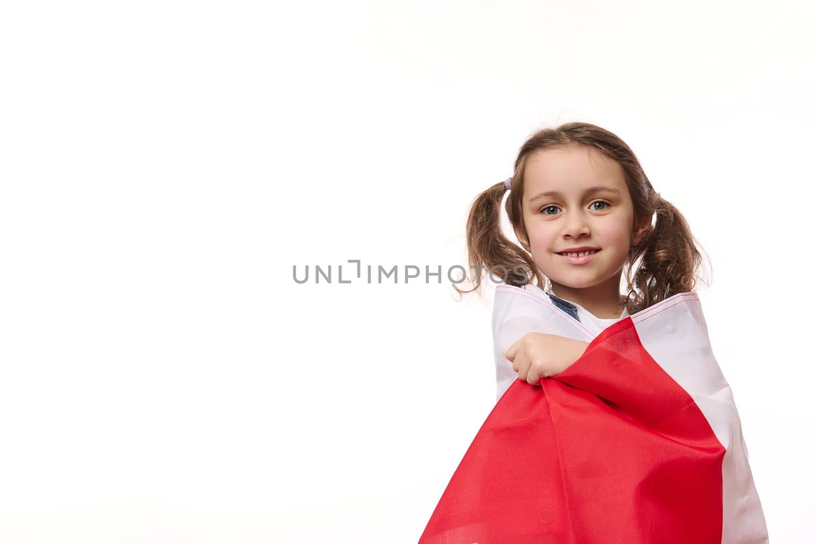 European little girl, proudly wrapping in flag of Poland or Canada, isolated on white background. Concept of national holidays. Patriotic events. Citizenships. Patriotism. Independence Day. Ad space