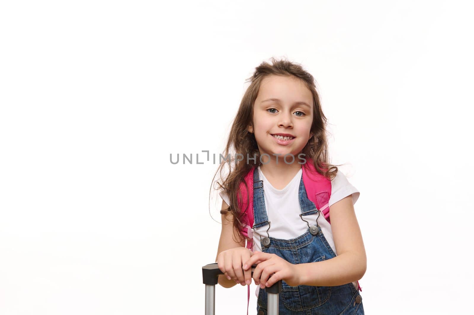 Caucasian adorable 5 year old traveler child, a little girl, dressed in denim overalls, with a pink backpack and suitcase, smiles sweetly looking at camera, isolated on white background. Free ad space