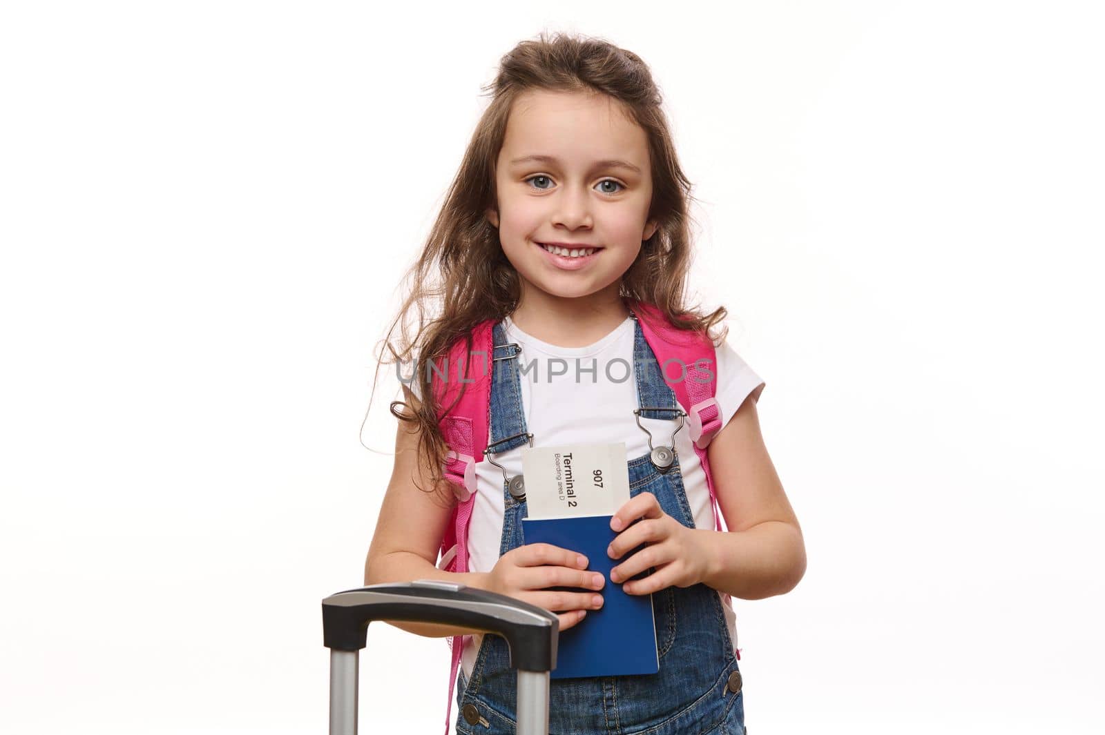 Lovely little girl in blue denim overalls, with a pink backpack, holds a boarding pass, smiles cutely looking at camera, by artgf