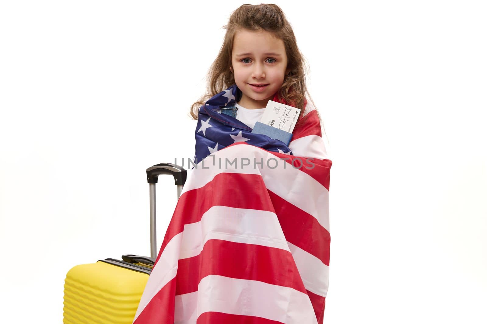 Lovely little girl wrapped in USA flag, holding air ticket and passport while standing with suitcase on white background with copy space for promotional text. Travel, immigration and tourism concept