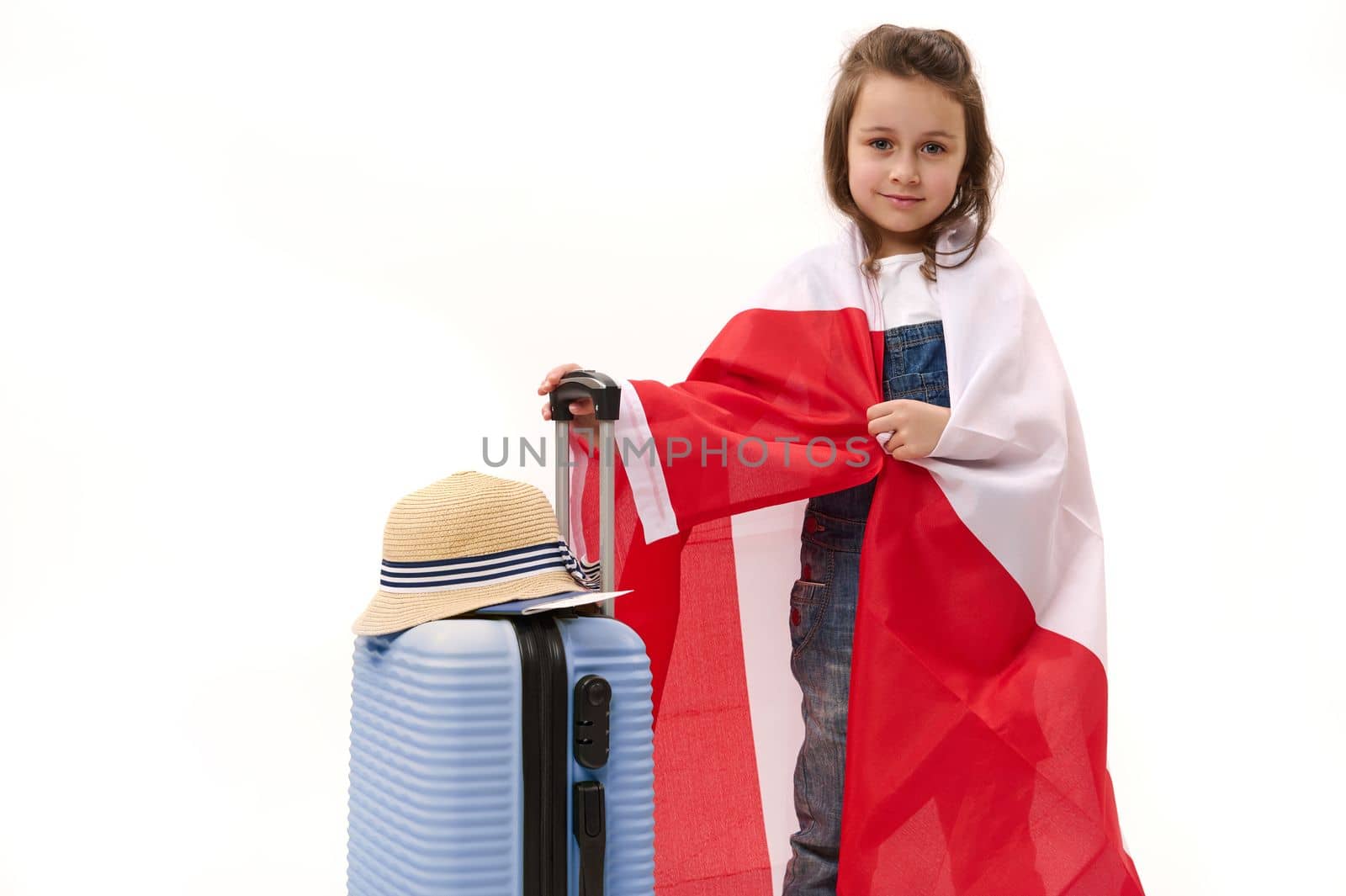 Caucasian cute little girl, little traveler, wrapping in Canada or Poland flag, smiles looking at camera, posing with blue suitcase, isolated over white background with free space for advertising text