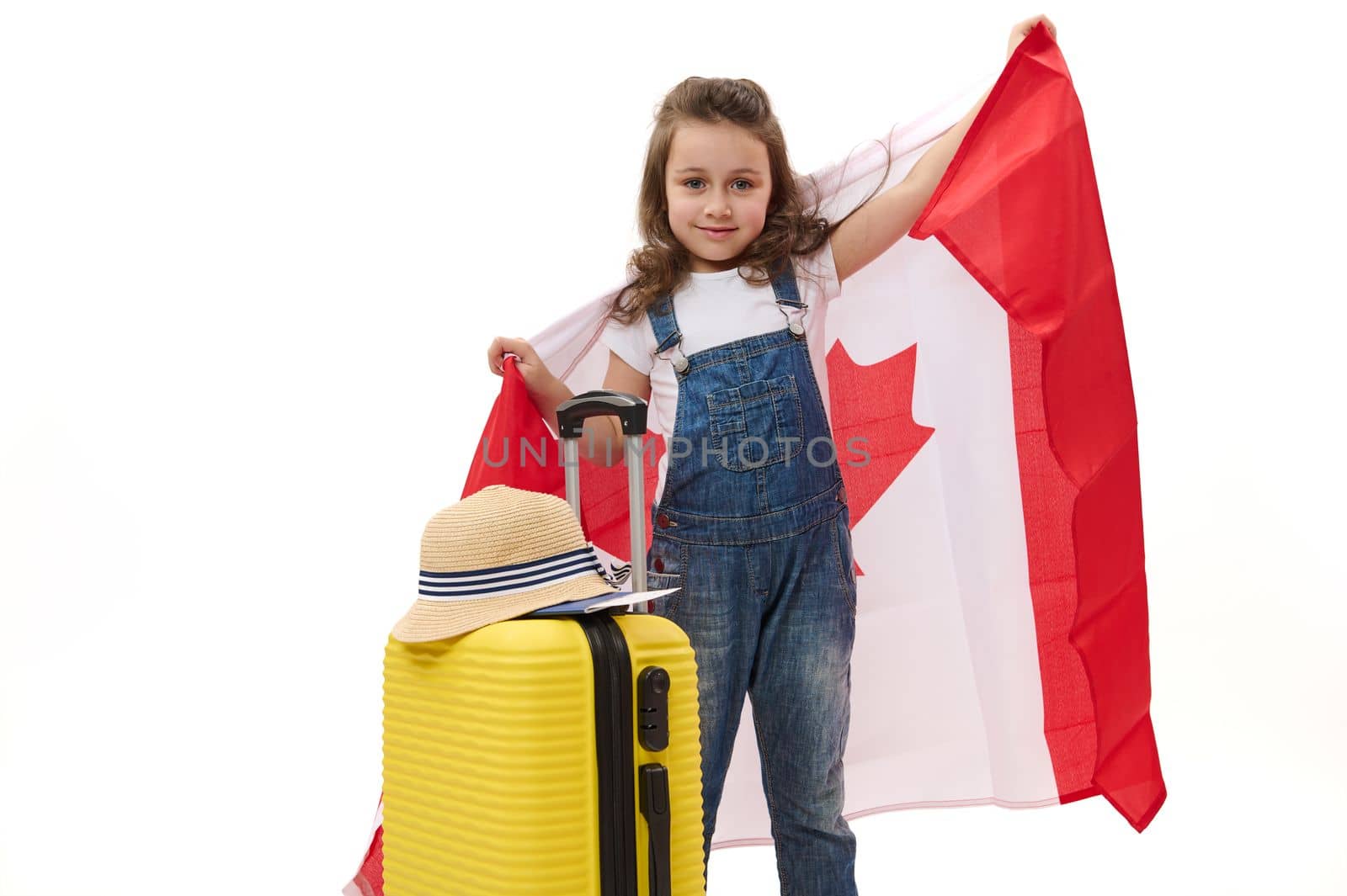 European little child preschooler girl carries Canadian flag, smiles looking at camera, posing with a suitcase over white background. Concept of study in Canada, emigration, learning foreign languages