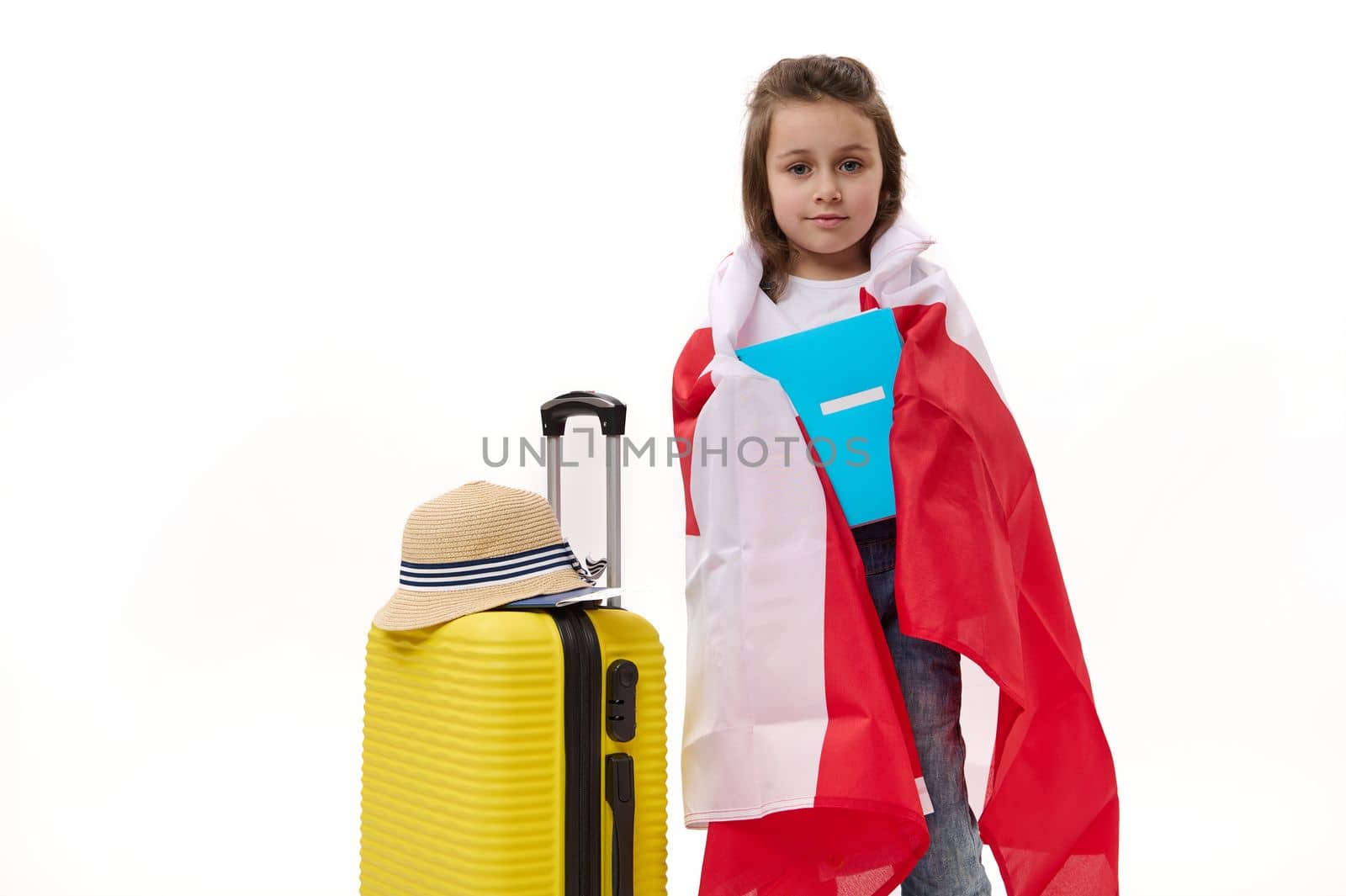 Happy first grader, Caucasian little child girl wrapping in Canadian flag, holding a copybook, posing with suitcase over white background. Concept of travelling to Canada for study. Language courses