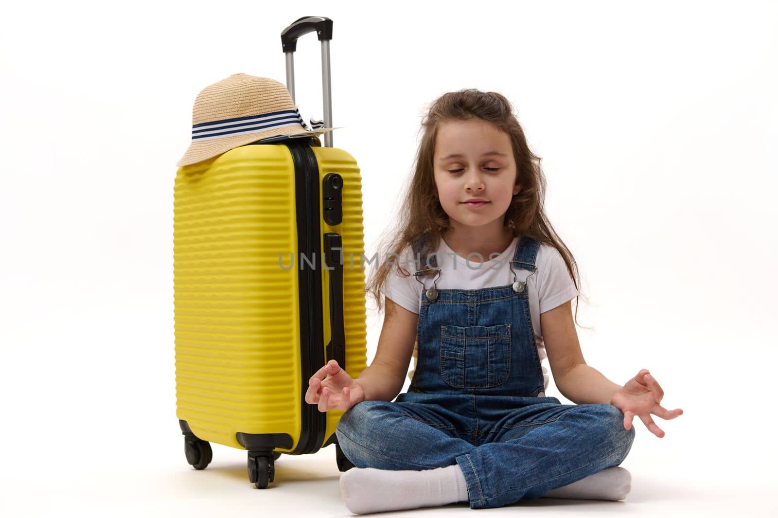 Peaceful little girl in denim overalls, enjoying meditation while waiting for her flight, sitting in lotus positions next to bright yellow suitcase, isolated over white background. Advertising space