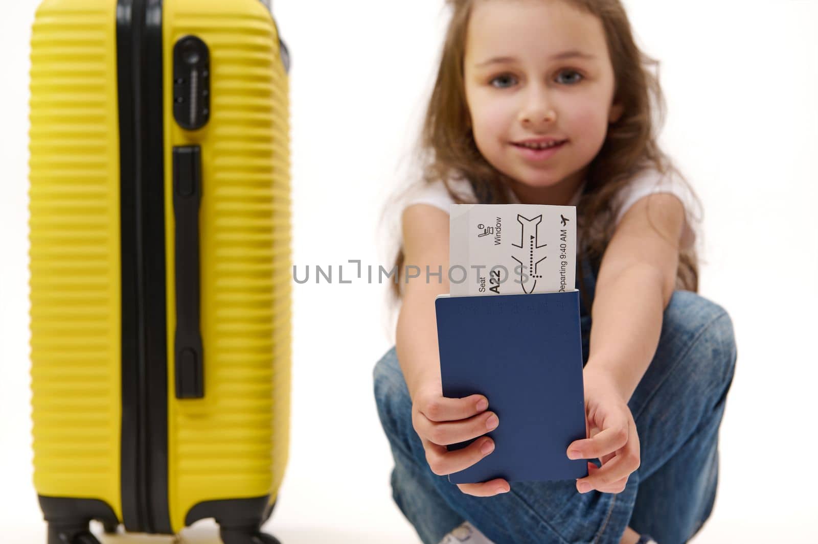 Details: boarding pass and identity documents for travel abroad in the hands of a blurred smiling little girl traveller with yellow suitcase, isolated over white background. Journey. Air flight trip