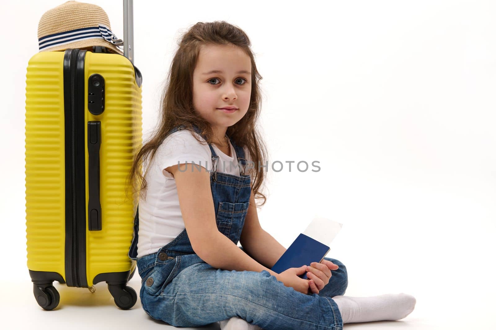 Adorable little girl traveler wearing blue denim overalls, with boarding pass, leaning on her yellow suitcase, smiling and looking at camera, isolated on white background. Travel Trip Tourism concept