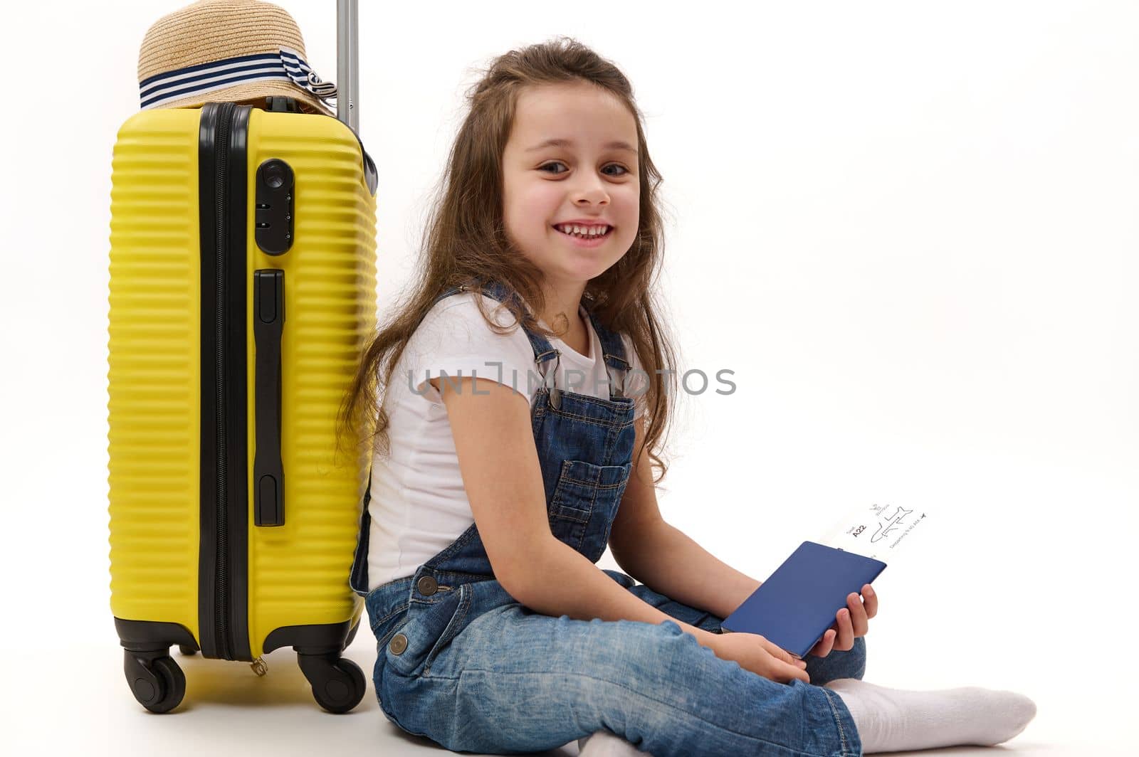 Adorable little girl passenger wearing denim overalls, sitting over white background with boarding pass in her hands, leaning on yellow suitcase and smiling a cheerful toothy smile, looking at camera