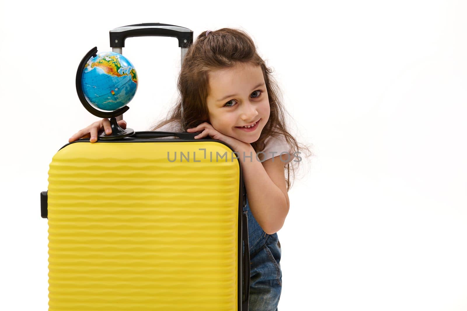 Close-up portrait of a beautiful little girl passenger with yellow suitcase and globe, cutely smiles looking at camera, isolated over white background. Travel. Tourism. Journey concept Free ad space
