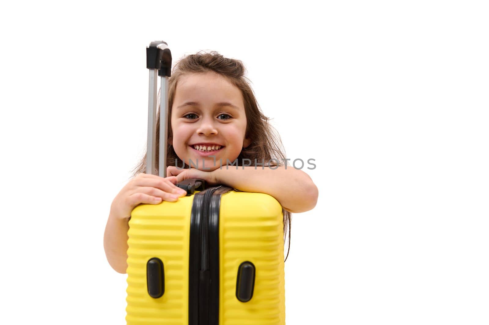 Mischievous little girl smiling a cheerful toothy smile looking at camera, leaning on a yellow suitcase. Travel concept by artgf