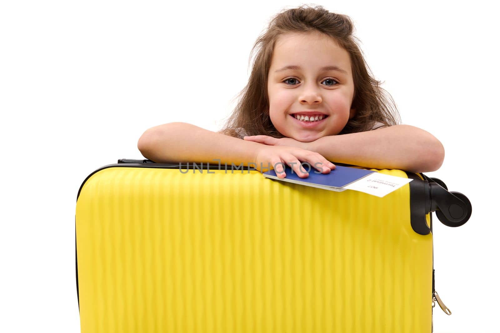 Adorable little child girl passenger with boarding pass, cutely smiles looking at camera, leaning on her yellow suitcase by artgf