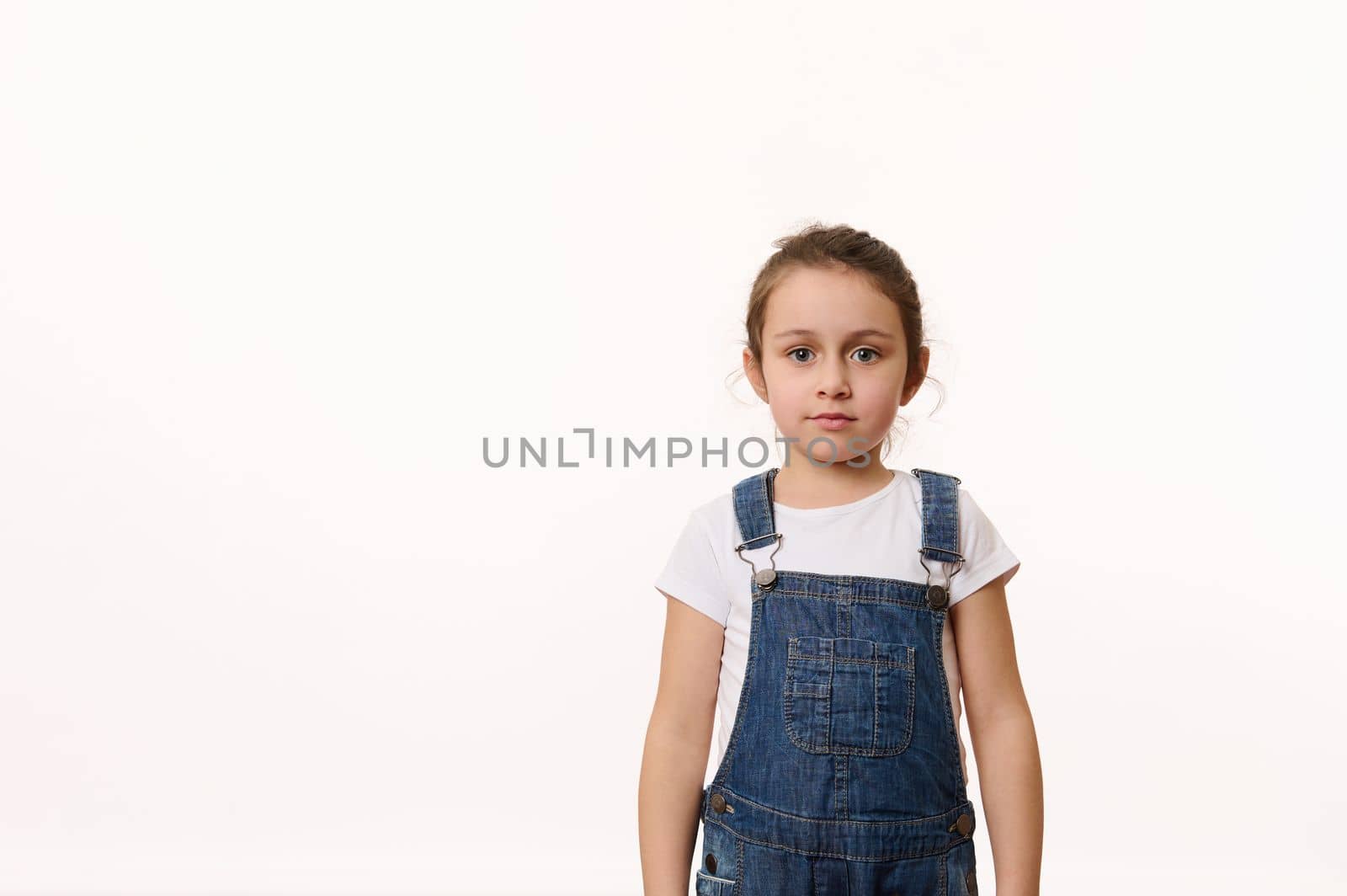 Confident portrait of an adorable Caucasian little child girl, wearing a white t-shirt and blue denim overalls, looking at camera, isolated over white background with copy space for promotional text.