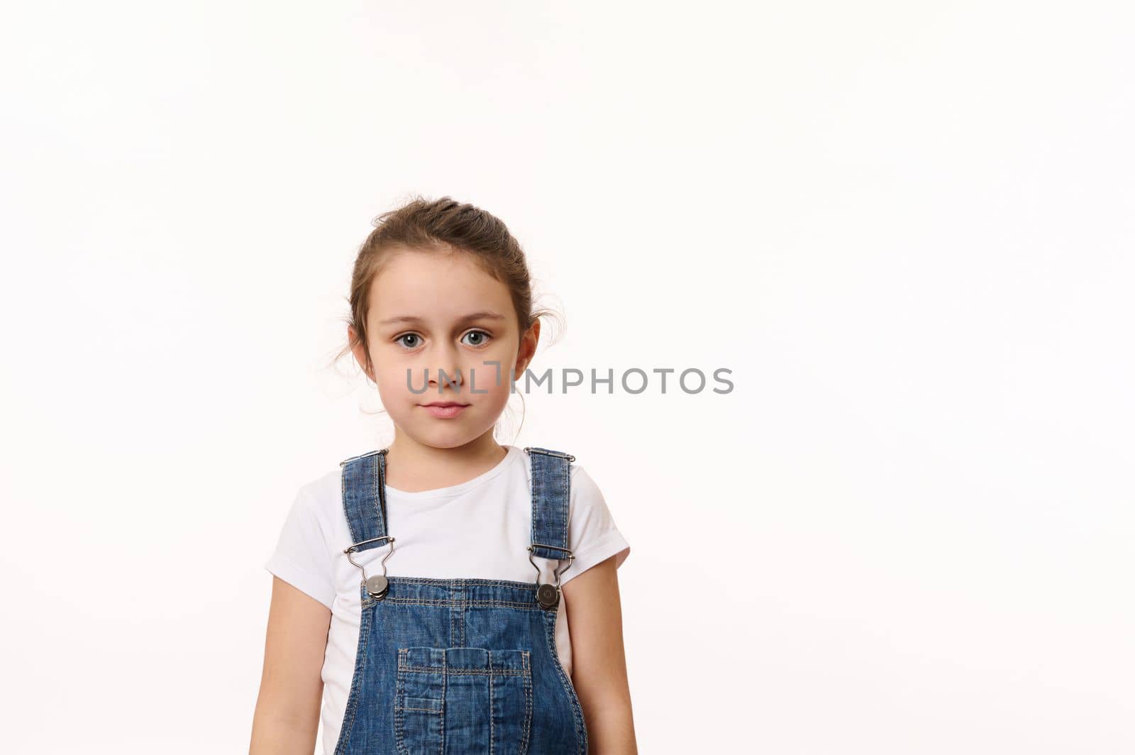 Isolated portrait on white background of a Caucasian adorable blue-eyed child, cute baby girl in white t-shirt and blue denim overalls, confidently looking at camera. Copy space for advertising text