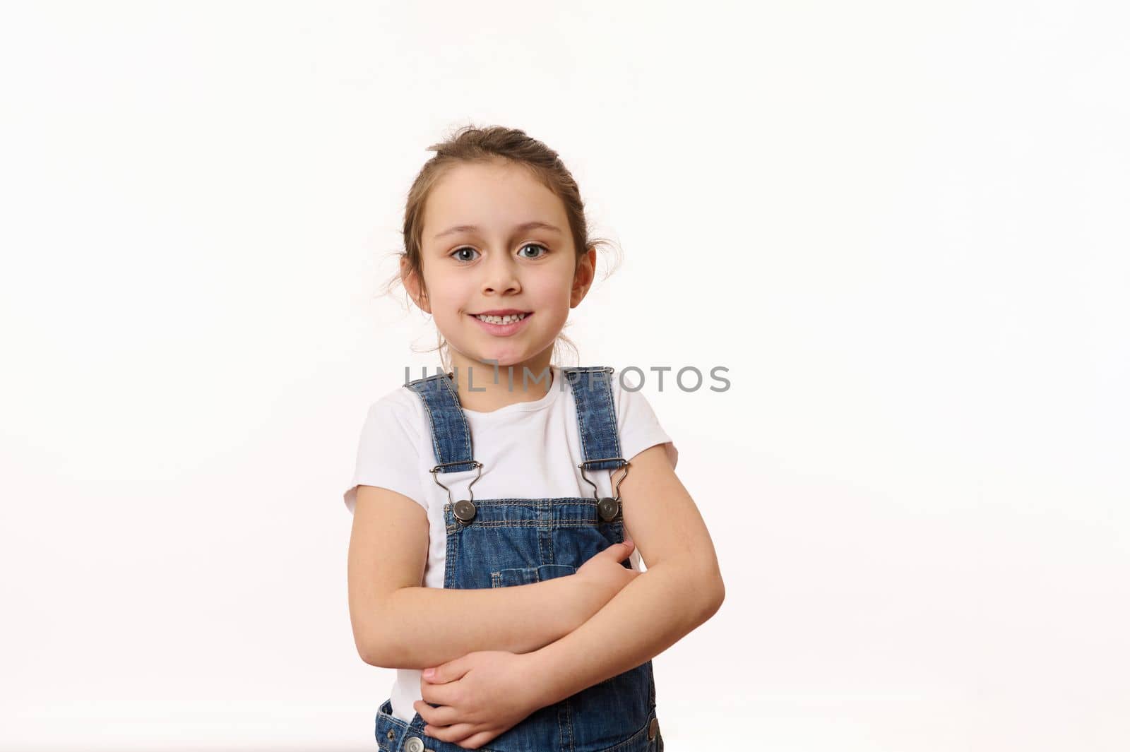 Emotional portrait of a Caucasian cute baby girl, wearing blue denim overalls, happily smiling looking at camera, hugging herself while posing over white background with copy ad space for promo text