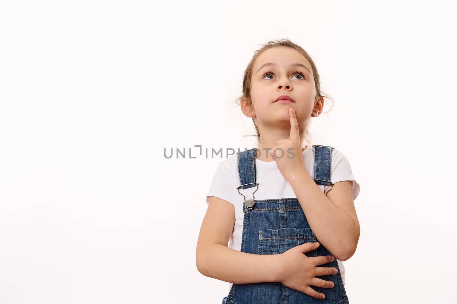 Beautiful pensive little girl wearing blue denim overalls, holding her finger on her chin, thoughtfully looking up at a copy advertising space for promotional text, isolated over white background