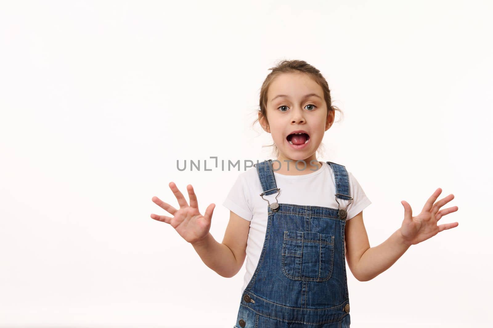 Emotional portrait on white background of a preschool child, overjoyed cute baby girl, wearing blue denim overalls, sweetly smiling, showing her hands palms to camera. Beautiful kids, happy childhood