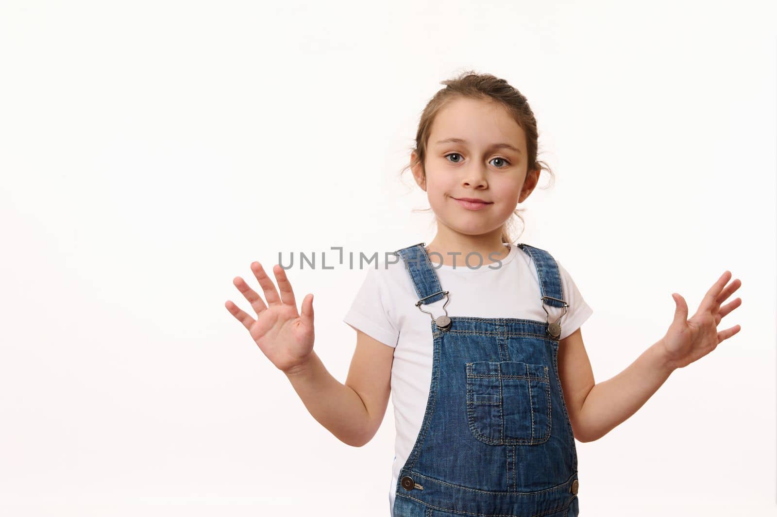 Emotional portrait on white background of beautiful preschool child, cute baby girl, wearing blue denim overalls, sweetly smiling, showing her hands palms to camera