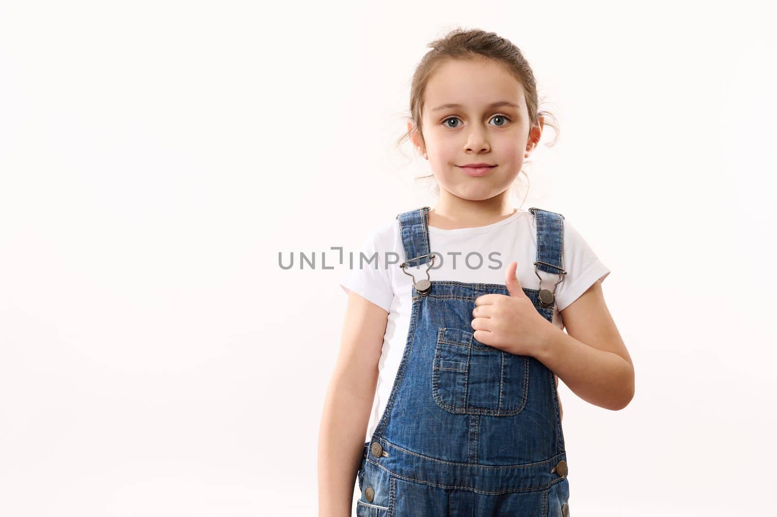 Beautiful European elementary age, preschool, first grader little child girl, wearing blue denim overalls, shows a thumb up, cutely smiles looking at camera, isolated over white background. Copy space