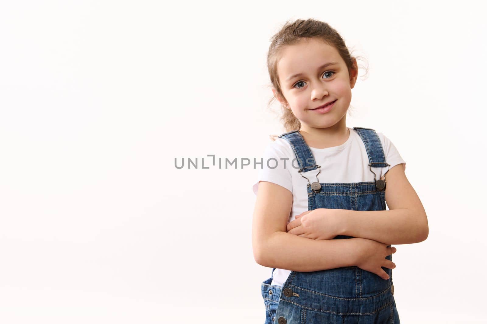 Happy cute adorable little child baby girl in blue denim overalls and white t-shirt, hugging herself, smiling toothy smile looking at camera, isolated over white background with copy advertising space