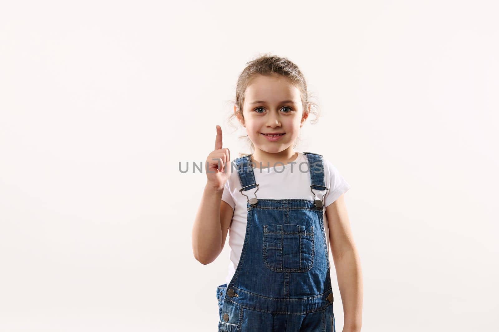 Isolated portrait on white background of a lovely little child girl, wearing blue denim overalls, pointing her finger up at a copy space for text on white background, cutely smiling looking at camera