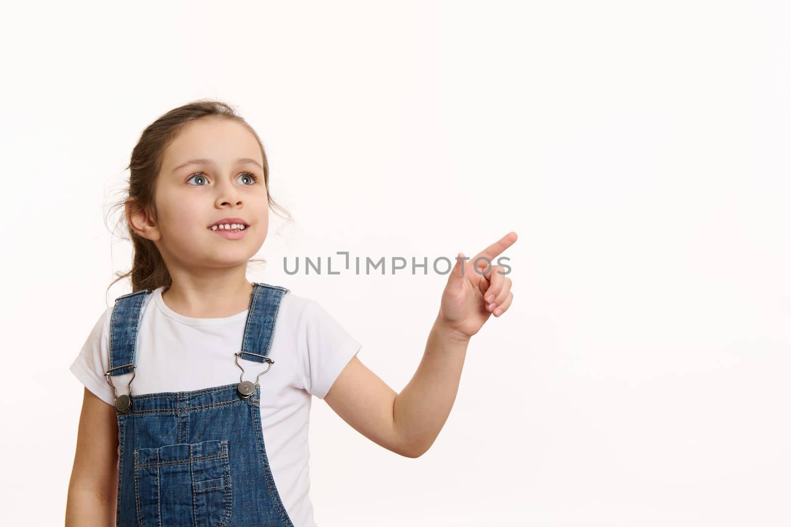 Horizontal studio shot on white background of a happy European little girl, adorable preschooler smart child, wearing blue denim overalls, pointing away on free space for your advertising content