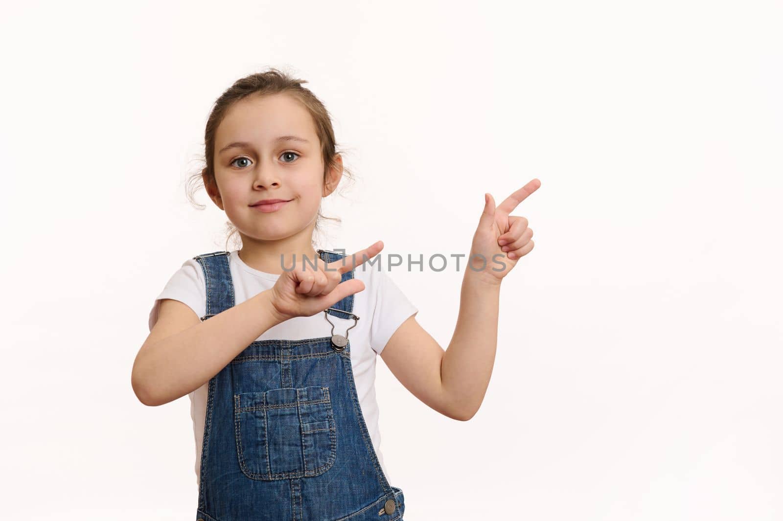 Isolated portrait on white background of a preschool Caucasian lovely child, adorable baby girl in blue denim overalls, pointing her finger at copy ad space, cutely smiling looking at the camera