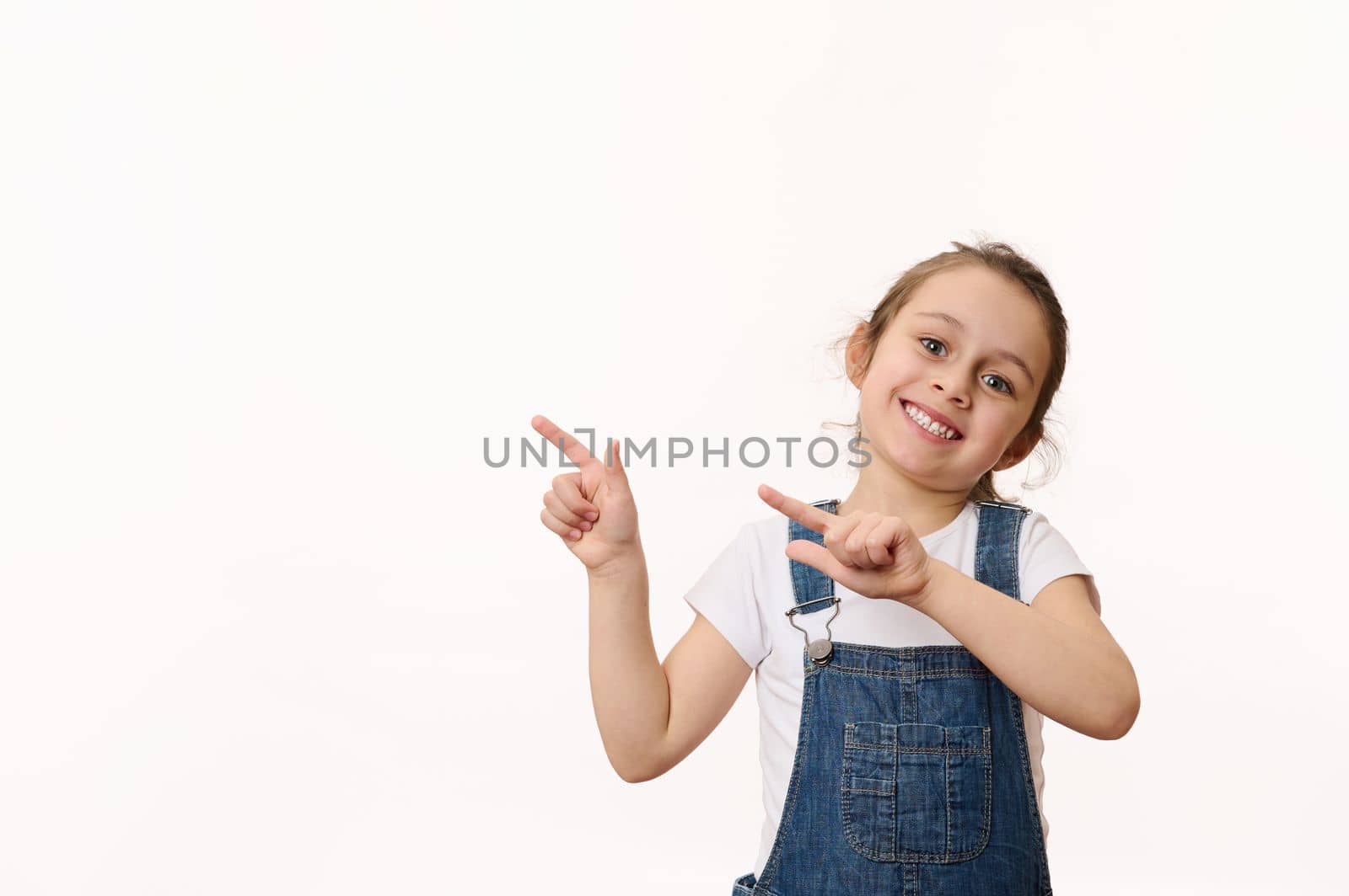 Adorable delightful Caucasian little child, a cute baby girl wearing a blue denim overalls, smiling a toothy smile, pointing at a copy space for promotional text, isolated over white background.