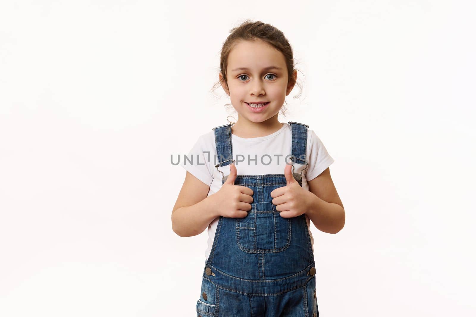 Adorable elementary age Caucasian little child, preschool kid, a lovely baby girl wearing blue denim overalls, shows thumbs up and sweetly smiles looking at camera, isolated on white background